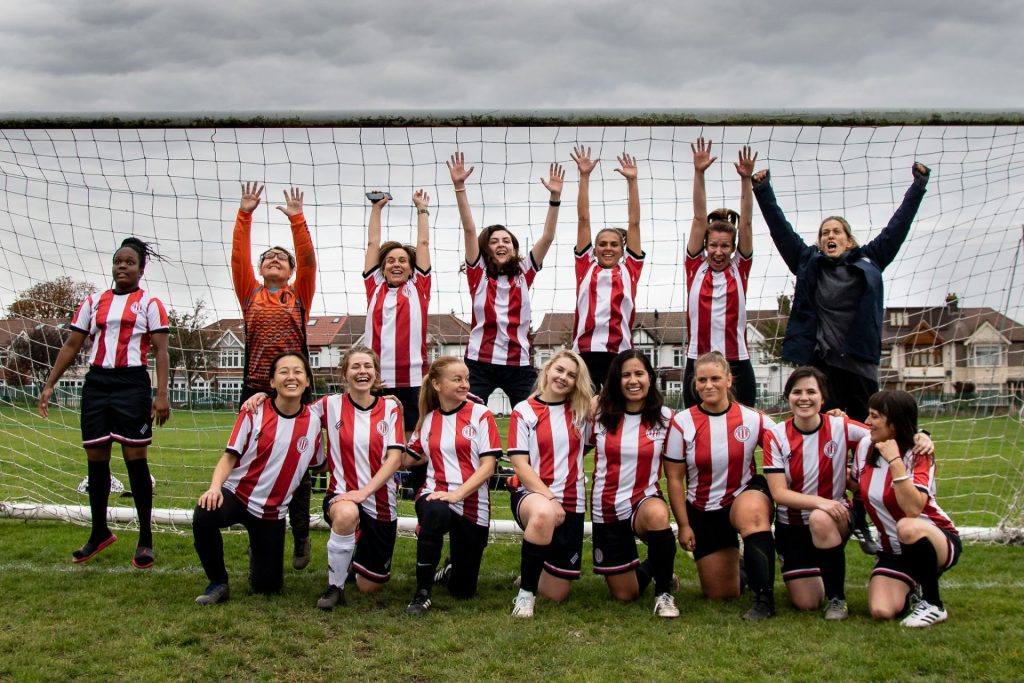 Clapton CFC Women's 11-aside beginners team pose together victoriously in a goal. Half of them kneel at the front, while the other half of the team jump for joy at the back. Image credit: Clapton CFC 
