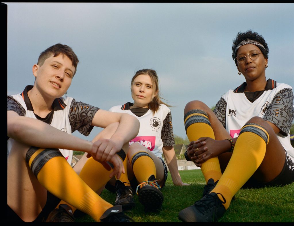 three players from Brockwell United sit on the pitch looking down at the camera. they are wearing their team colours of yellow, white and black