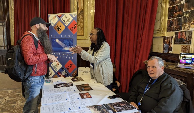 An attendee at the job fair stands in conversation with a representative from Delfont Mackintosh theatres, either side of a table, with a third person sat in the foreground.