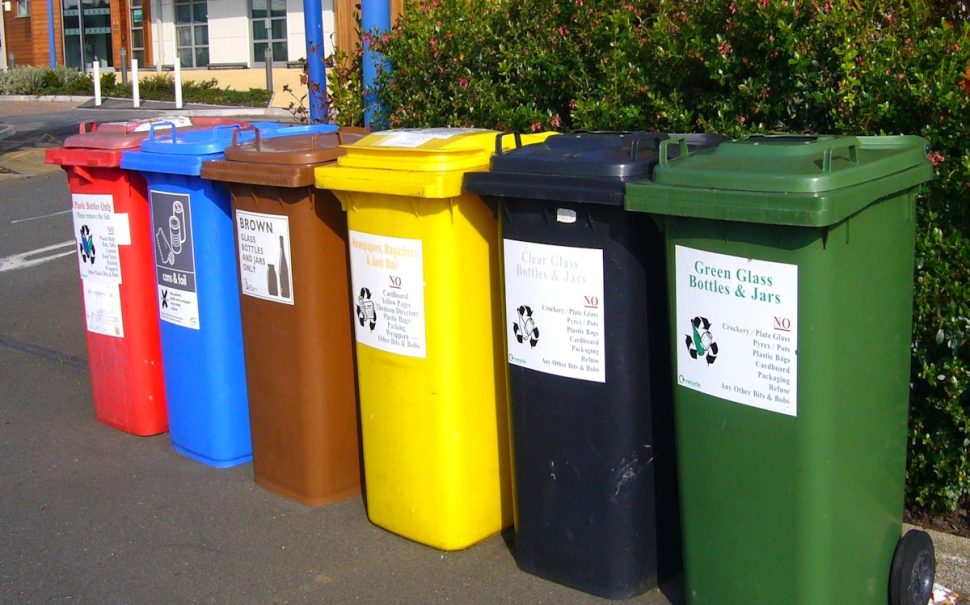 Six different types of bins, including five recycling and one rubbish, are sitting lined up on the side of the street