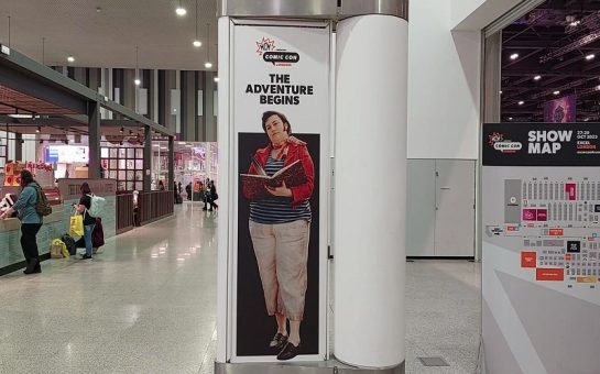 A sign displaying a man in cosplay outside of the main exhibition hall in ExCel London. The caption reads 'The Adventure Begins'.