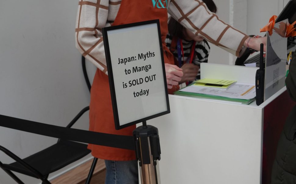 Lady stands at the sales counter at the Young V&A. In front of her is a sign saying Japan: Myths to Manga is sold out.