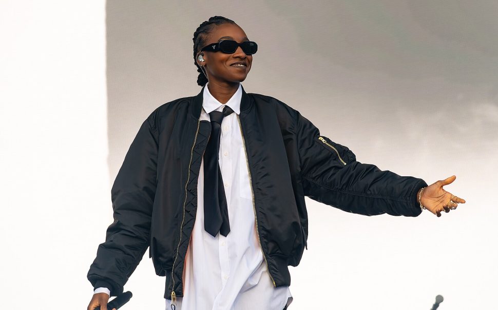 Rapper Little Simz in black sunglasses, black skirt and tie. She is wearing a bomber jacket.