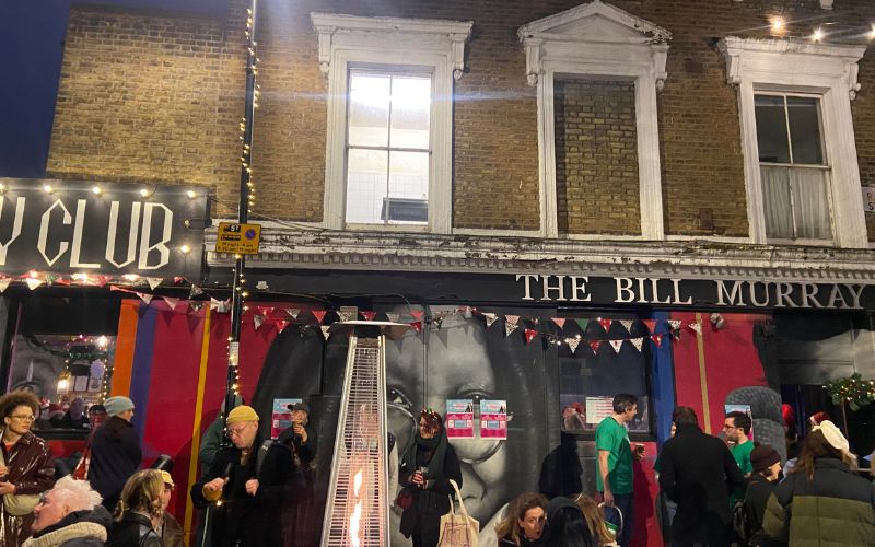 exterior of the Bill Murray pub. People milling about outside, by an outdoor flame. The wall has a mural of Whoopie Goldberg and posters up for the fundraiser with QR codes to donate with.