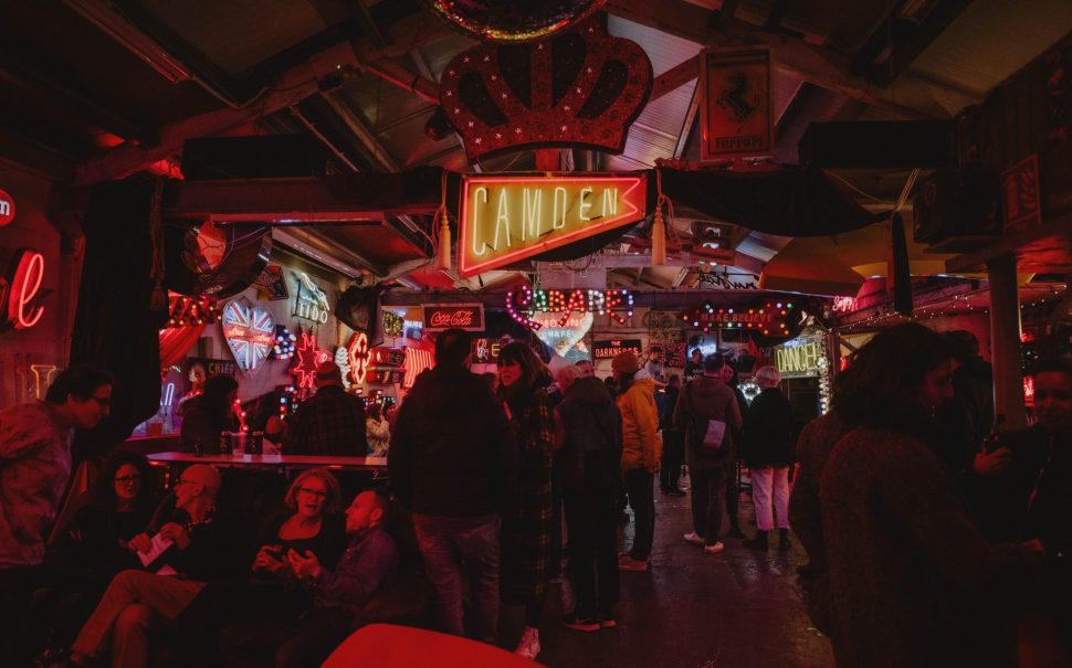 People standing inside a darkly-lit venue with a neon Camden sign hanging from above.