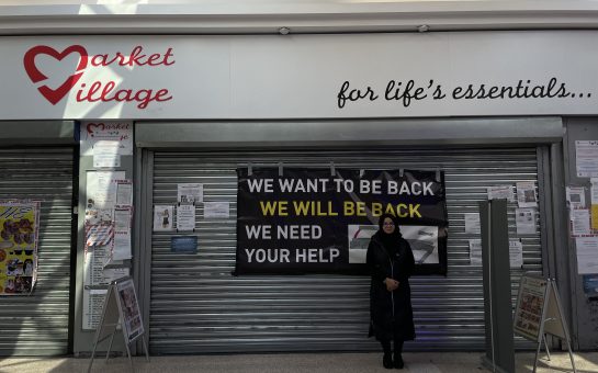 Shop manager Farhana Rasheed stands in front of market village with sign behind saying 'we want to be back, we will be back, we need your help.'