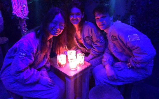Three people sitting round a small, oblong table smile up at the camera. The table has four luminous cocktails atop it. The people are bathed in a purple light.
