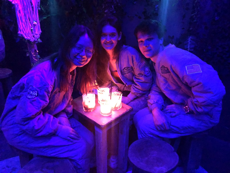 Three people sitting round a small, oblong table smile up at the camera. The table has four luminous cocktails atop it. The people are bathed in a purple light.