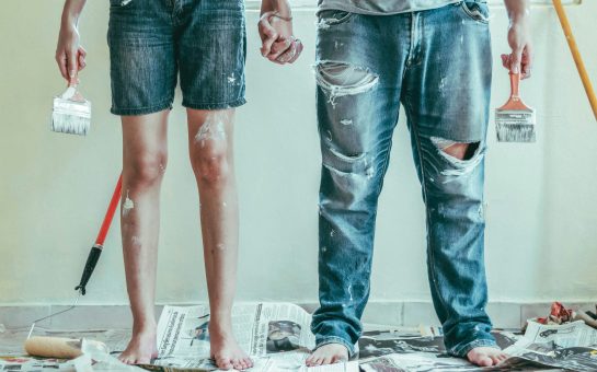 Two people wearing denim trousers and shorts are holding hands. You can only see their legs. They are bare foot standing on newspaper and there is paint on their clothes.