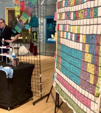 Image of a stall at the "Lates" event. On the left-hand side of the image, a group of five people can be seen near a black stall that is decorated with science crafts. One member of the group is pointing towards the right-hand side of the image where there is a wooly periodic table hanging up.