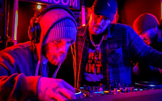 Two DJs stand at a set of decks in a nightclub.
