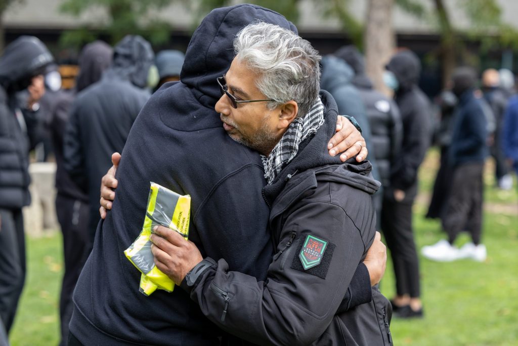 Two men embrace, one wearing black with his hood up and the other of Bengali heritage with grey hair. He is wearing a kaffiyeh, holding a high vis vest and displaying a Bengali regiment insignia on his sleeve.