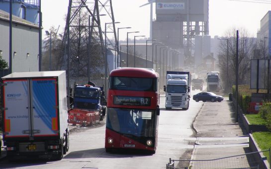 EL2 London red double decker bus driving through dusty dagenham dock industrial site with other lorries and windmills in the background
