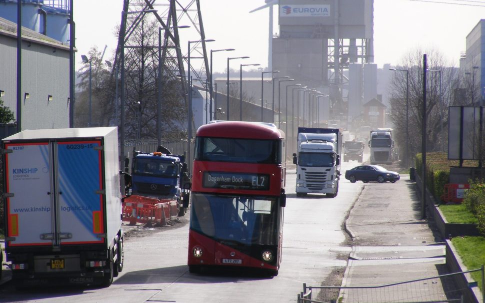 EL2 London red double decker bus driving through dusty dagenham dock industrial site with other lorries and windmills in the background