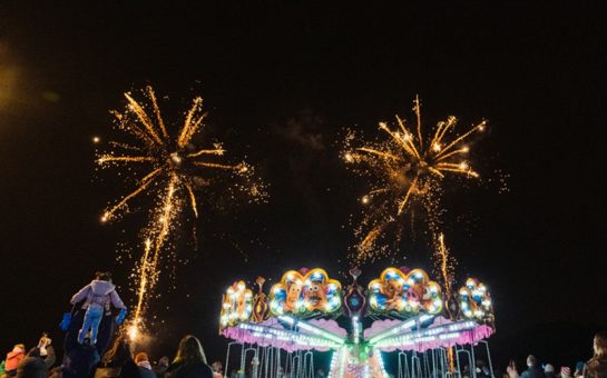 White fireworks exploding against dark night sky, with brightly lit carousel in the foreground.