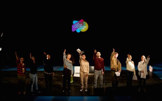 A photo of the Arts Council England funded Squint Playwriting Award, with nine young writers on a stage gesturing towards the sky
