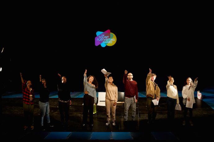 A photo of the Arts Council England funded Squint Playwriting Award, with nine young writers on a stage gesturing towards the sky