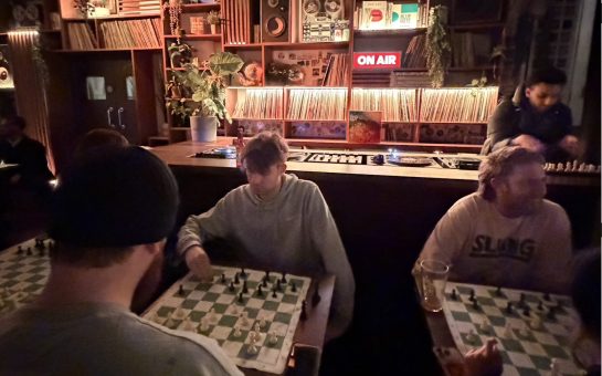 Two tables of couples playing chess. Shelves of records and DJ decks in the background.