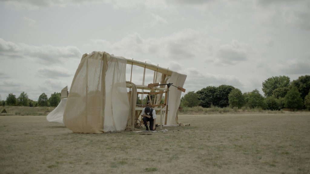 A still image showing a man kneeling in Hackney Marshes with a cream sheet behind him, from the Arts Council funded 900 Feet Up by Odd Eyes Theatre