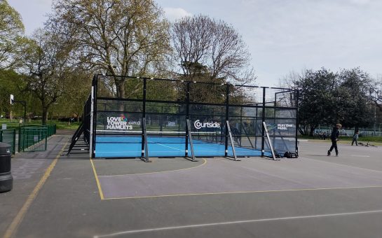 A padel court with blue astroturf and black fenced walls, Green Gardens, with basketball courts in the foreground and grey tarmac