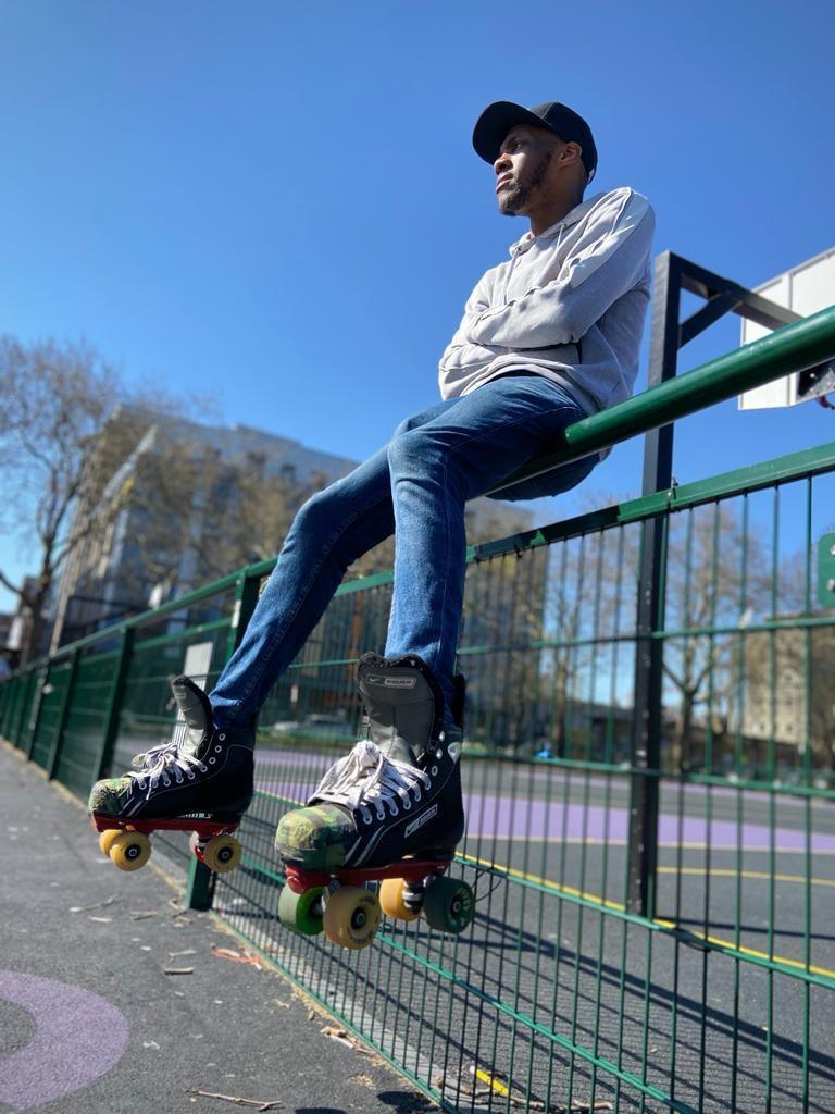 Skater Jamie Simon photographed sitting on the side fence at Bethnal Green Gardens, wearing a grey hoodie, jeans and roller skates, looking off into the distance