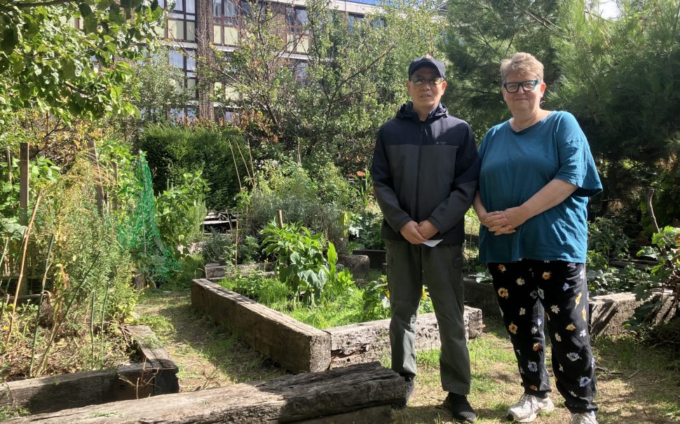 Xing Wang and Angela Large stand side-by-side next to seedbeds under the shade of mature trees, the St John’s Estate behind them.