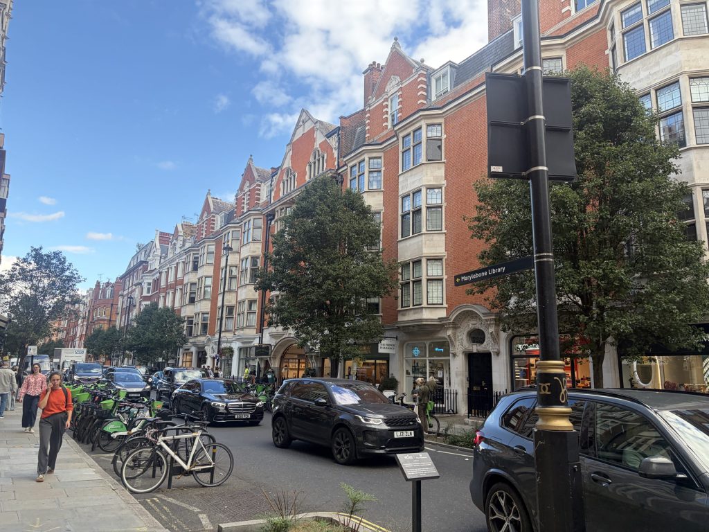Rush-hour traffic on New Cavendish Street in Marylebone, with cars, bicycles and pedestrians outside Marylebone Library