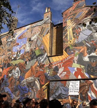 Marchers pass a 15m x 20m mural depicting the Battle of Cable Street, a historic clash between police, fascist protestors, and counter protestors in London’s East End. A woman holds up a sign in front of the mural reading “I believe that one fine day, I believe that one fine day, the power of love will rise above the love of power.”