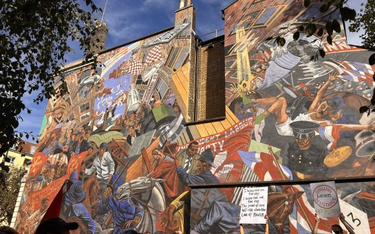 Marchers pass a 15m x 20m mural depicting the Battle of Cable Street, a historic clash between police, fascist protestors, and counter protestors in London’s East End. A woman holds up a sign in front of the mural reading “I believe that one fine day, I believe that one fine day, the power of love will rise above the love of power.”