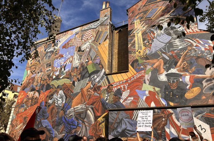 Marchers pass a 15m x 20m mural depicting the Battle of Cable Street, a historic clash between police, fascist protestors, and counter protestors in London’s East End. A woman holds up a sign in front of the mural reading “I believe that one fine day, I believe that one fine day, the power of love will rise above the love of power.”