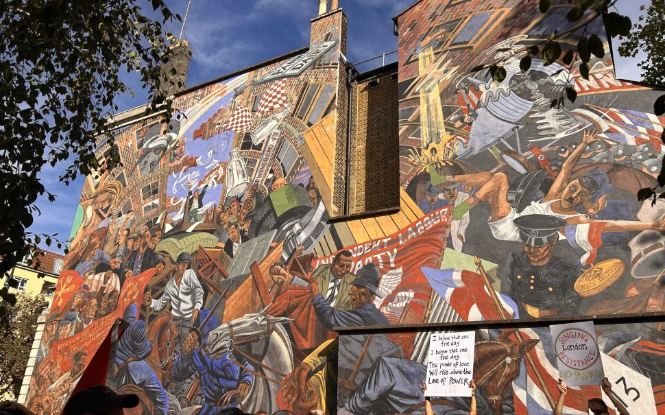 Marchers pass a 15m x 20m mural depicting the Battle of Cable Street, a historic clash between police, fascist protestors, and counter protestors in London’s East End. A woman holds up a sign in front of the mural reading “I believe that one fine day, I believe that one fine day, the power of love will rise above the love of power.”