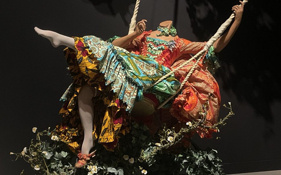 An image of The Swing by Yinka Shonibare, a headless mannequin in a brightly coloured patterned dress, on a swing surrounded by oak twigs and artificial foliage.